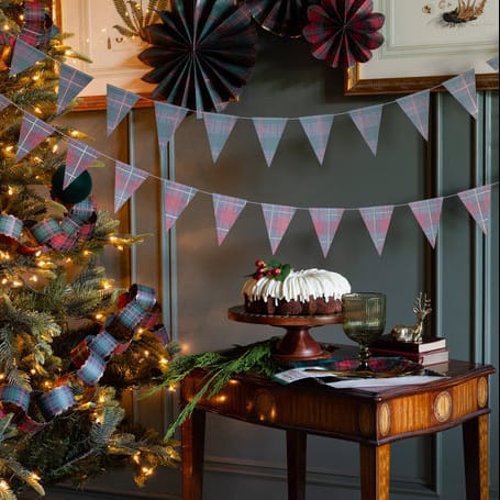 A cozy holiday scene with a decorated Christmas tree on the left, featuring warm white lights and plaid ribbon garlands. A small wooden table on the right holds a white-frosted cake on a stand, a glass, books, and festive papers against a dark gray paneled wall.