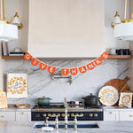 A styled kitchen interior with a marble backsplash and island, featuring a decorative orange banner that reads 'GIVE THANKS' hung above the stove. White cabinets, gold hardware, pendant lights, and Thanksgiving-themed dishware are visible.