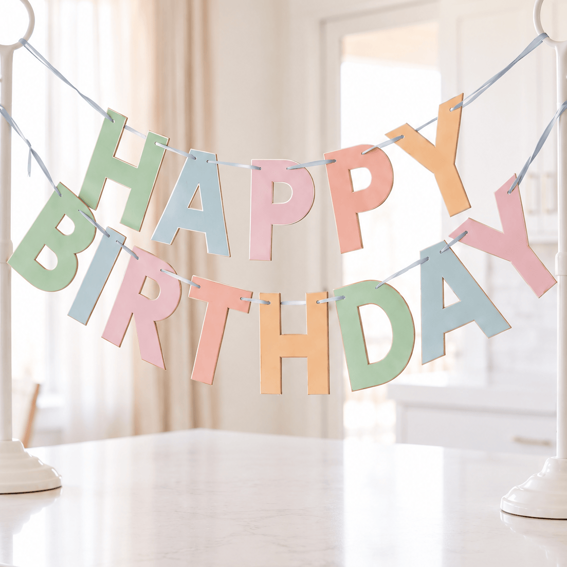 A pastel 'HAPPY BIRTHDAY' banner made of hanging letters displayed on a kitchen island, with two white stand holders in a bright, modern kitchen setting.