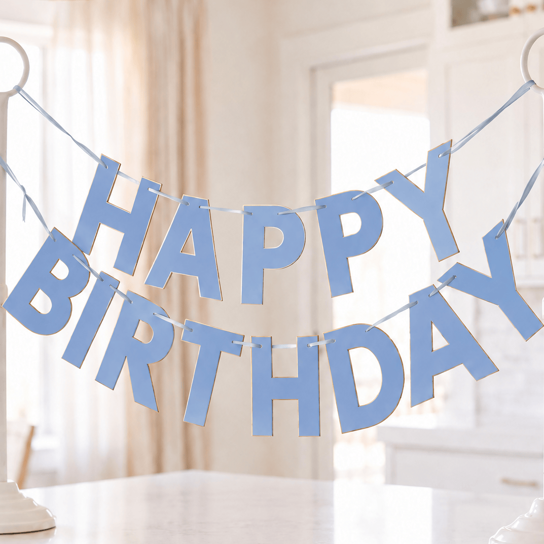 A light blue 'HAPPY BIRTHDAY' banner made of paper cutout letters, displayed on two white stand holders on a white marble kitchen island with a blurred kitchen background.