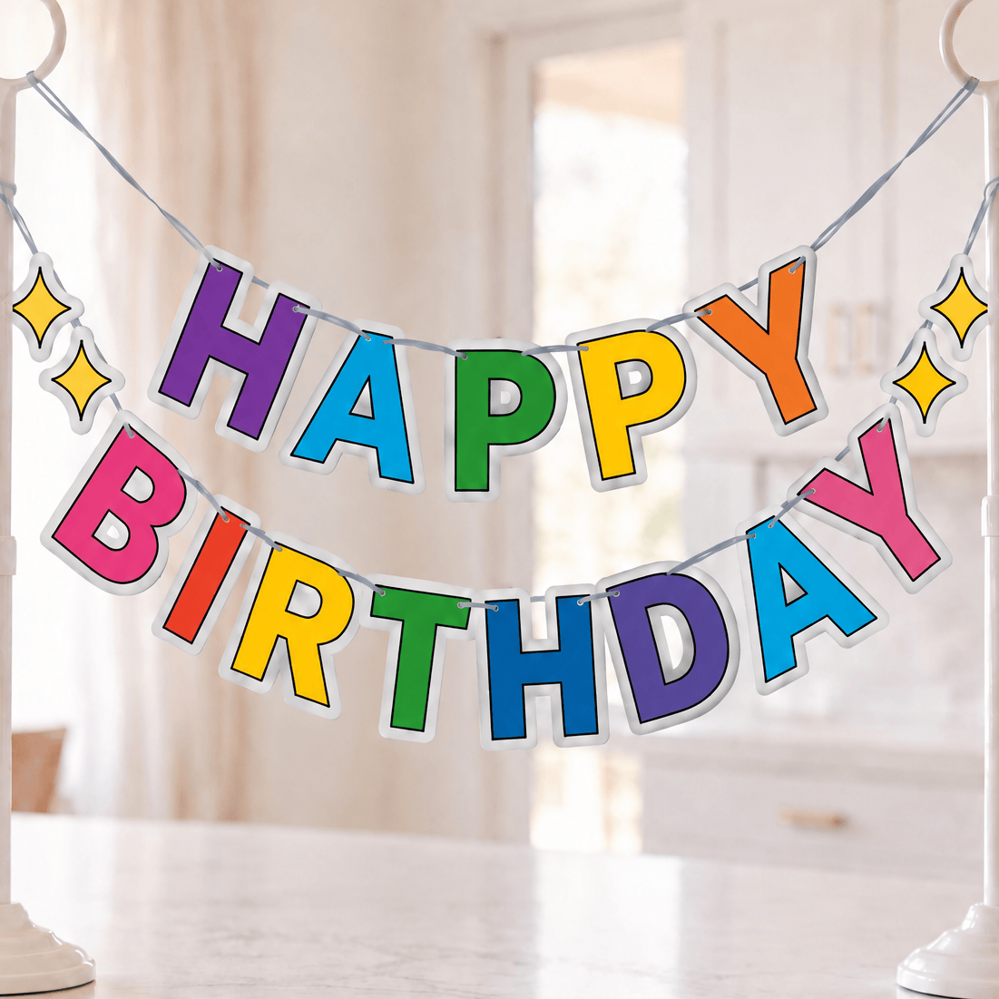 A colorful 'HAPPY BIRTHDAY' banner with raised letters in rainbow colors, suspended between two white floor stands on a kitchen island. The background shows a softly blurred bright kitchen interior with pendant lights and cabinets.