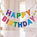 A colorful 'HAPPY BIRTHDAY' banner with raised letters in rainbow colors, suspended between two white floor stands on a kitchen island. The background shows a softly blurred bright kitchen interior with pendant lights and cabinets.