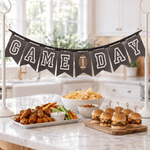 A 'GAME DAY' banner made of black pennants with white block letters and a small football detail hangs on a kitchen island. The kitchen is styled with football snacks including chicken wings, chips, salsa, and sliders on a marble countertop.